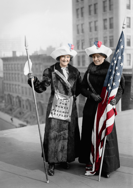 Black and white vintage photo of two women suffragettes with a bag that says Vote for Women. Holding an American Flag that is colorized.